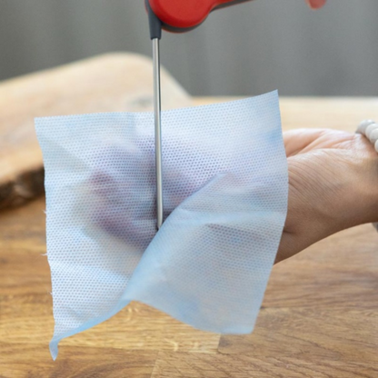 Une main tient délicatement un tissu bleu, avec des ciseaux prêts à couper sur une surface en bois, mettant en valeur la précision rappelant l'artisanat Lingette pour sondes - 6 cuves x 180 par Thermometre.fr.