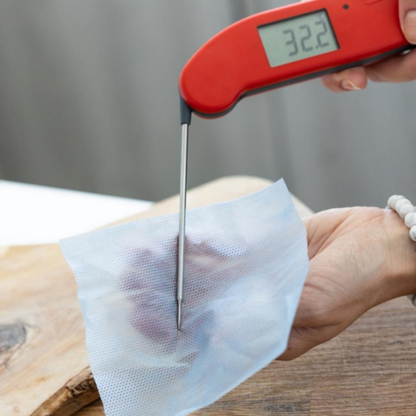 Une main utilise une Lingette pour sondes Thermometre.fr pour mesurer la température à travers un tissu en maille bleue recouvrant un morceau de viande, affichant 32,2°C, garantissant la sécurité contre Listeria et Salmonella.