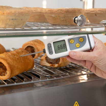 Une main tient un Thermomètre intelligent TempTest 1 de Thermomètre.fr, affichant 66,8°C, pour mesurer la température rapide d'une pâtisserie cuite sur une grille métallique.