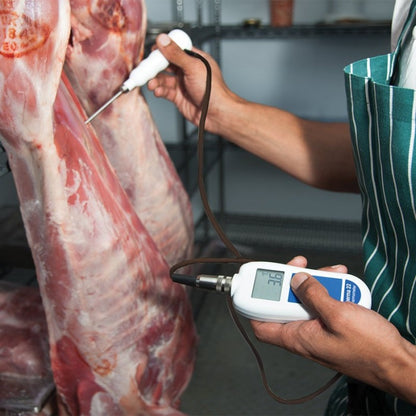 Une personne portant un tablier rayé utilise un thermocouple Therma 22 de Thermomètre.fr pour mesurer la température d'une viande suspendue dans un local de stockage.