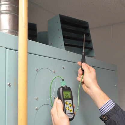 Une personne tient un thermomètre numérique industriel Therma Elite de Thermomètre.fr, avec un fil vert connecté à un port d'un appareil CVC de couleur sarcelle, vérifiant la température affichée à 20,4°C sur son écran LCD rétroéclairé.