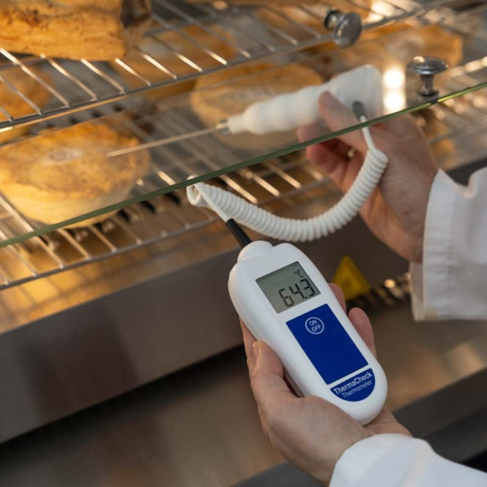 Personne utilisant le ThermaCheck avec sonde alimentaire de Thermomètre.fr pour vérifier la température des aliments à l'intérieur d'un four, l'appareil affichant 64,3°C. Précision assurée pour une cuisson parfaite à chaque fois.