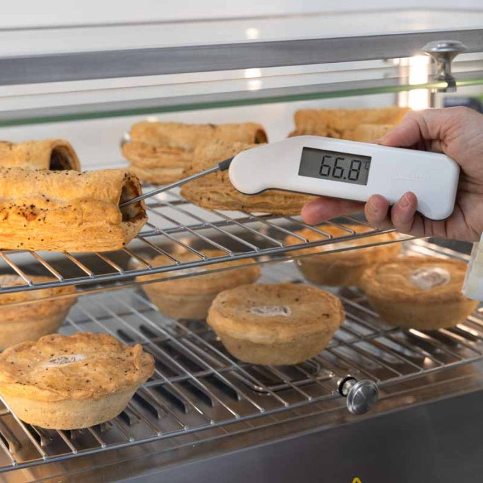 Une main utilise un Thermomètres Thermapen® Classic de Thermomètre.fr pour mesurer la température précise (66,8) d'un rouleau de saucisse sur une grille de four aux côtés d'autres tartes.