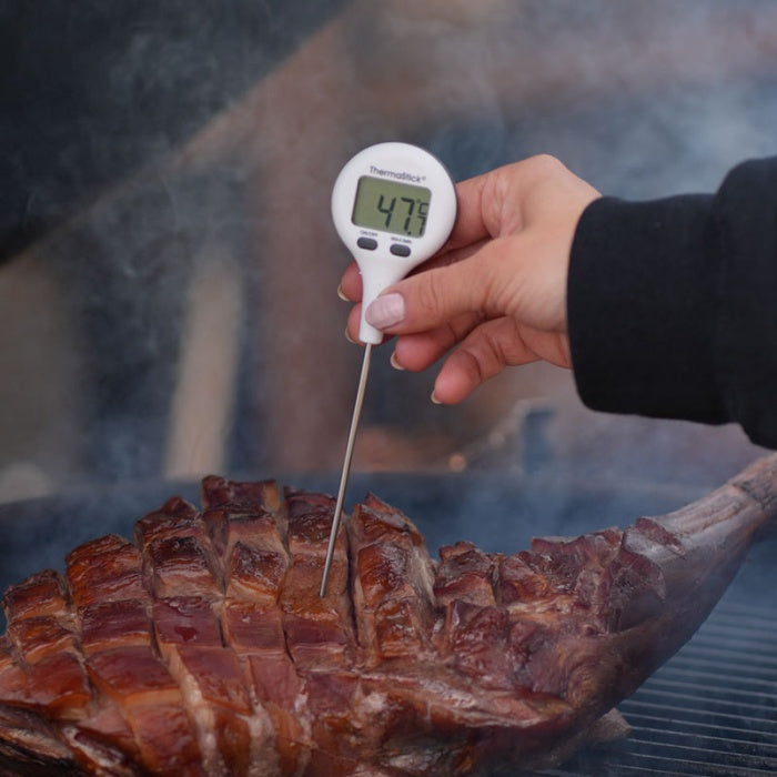 Une main tenant un Thermomètres de poche ThermaStick de Thermomètre.fr avec un écran LCD facile à lire affichant 47,9°C, inséré dans un gros morceau de viande cuit et tranché sur un grill.