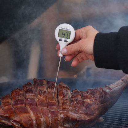 Une main tenant un Thermomètres de poche ThermaStick de Thermomètre.fr avec un écran LCD facile à lire affichant 47,9°C, inséré dans un gros morceau de viande cuit et tranché sur un grill.